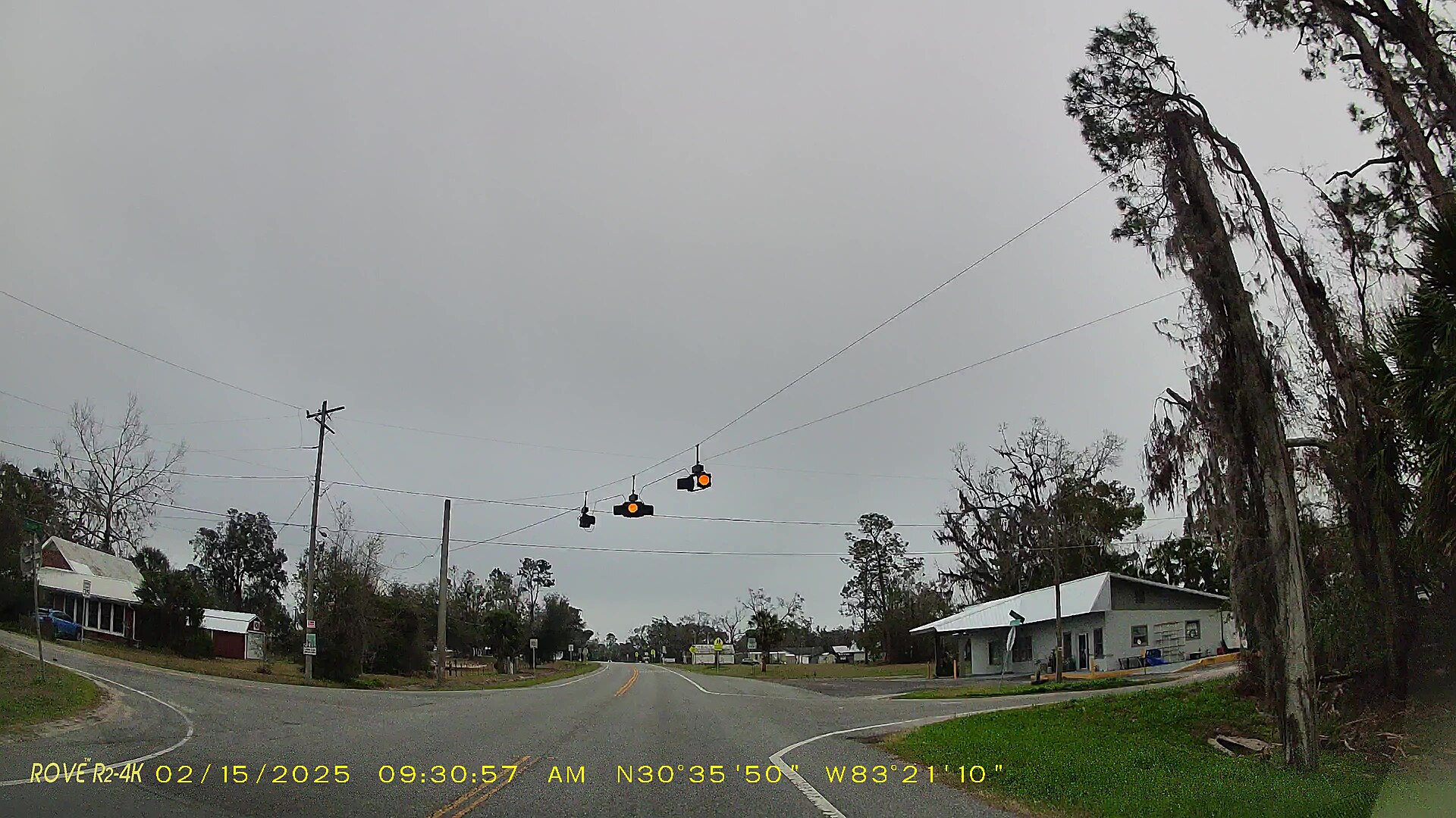 Dashcam view of a rural intersection with flashing amber signals under a grey sky